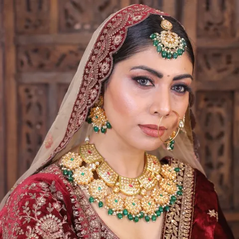 A close portrait of a bride in traditional makeup and gold jewellery, wearing a red and gold embroidered dupatta and outfit, against a carved wooden backdrop.